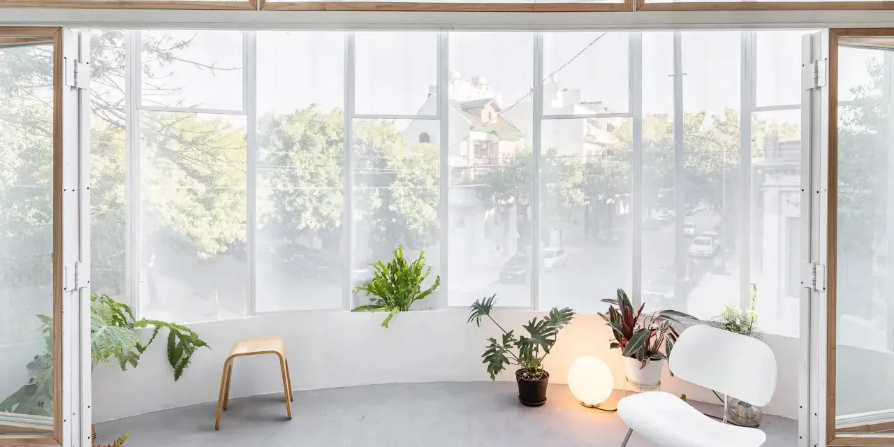 Interno luminoso con piante, sedia bianca e sgabello in legno, affacciato su una strada alberata da una veranda balcone.
