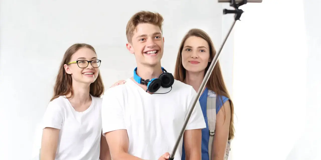 Trzech przyjaciół robi selfie na kijku. Chłopak z przodu uśmiecha się szeroko, dziewczyna w okularach po lewej stronie też, a ta z tyłu patrzy w obiektyw. To świetny sposób, jak robic dobre zdjecia!