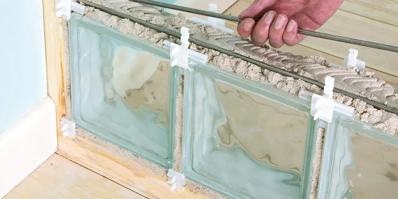 Pose de brique de verre extérieur : une main insère des barres de renfort dans le mortier entre les briques de verre, en cours d'installation.