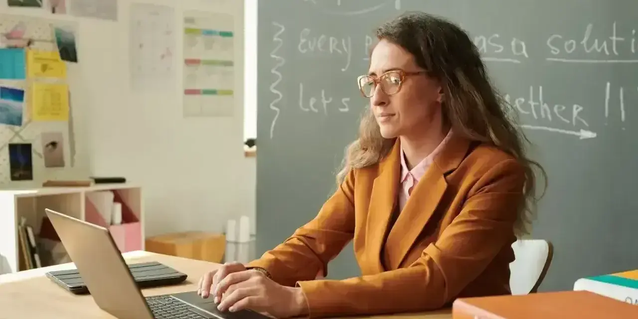 Nauczyciel początkujący w okularach pracuje przy laptopie w klasie. Na tablicy napisy po angielsku.