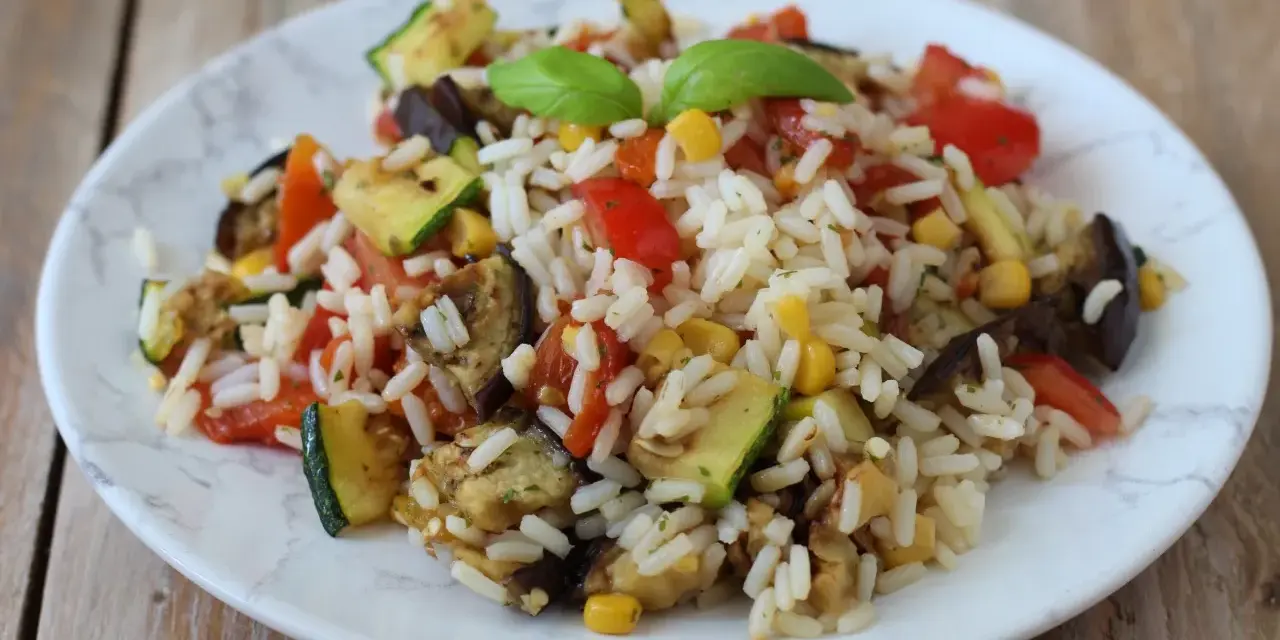 Un piatto colorato di insalata di riso vegetariana con zucchine, melanzane, pomodori e mais, guarnito con basilico fresco.