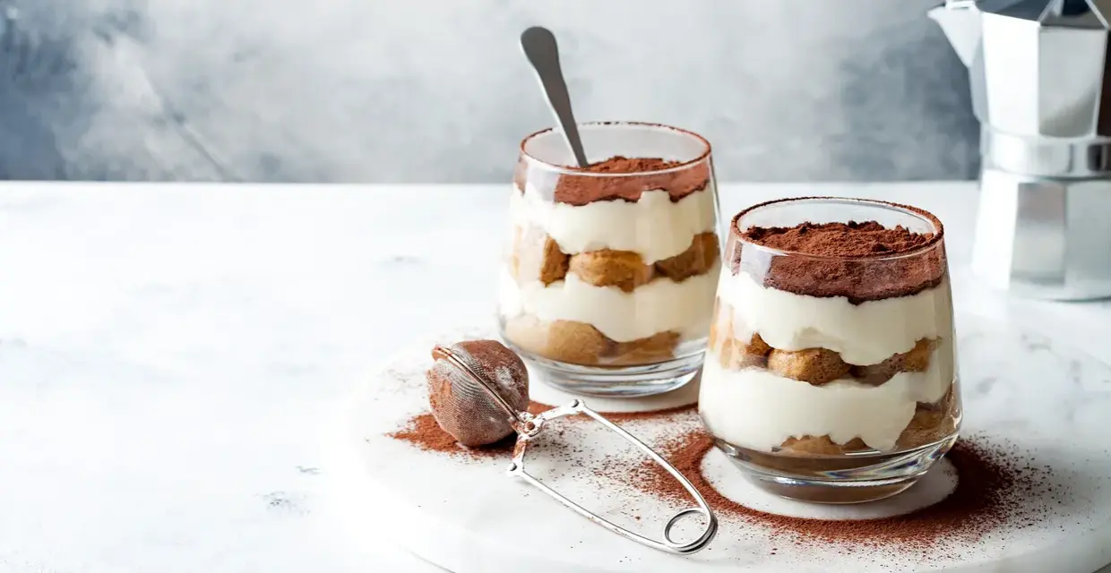 Dwa pucharki z kremowym tiramisu bez jajek, posypane kakao. Obok sitko do kakao i kawiarka.