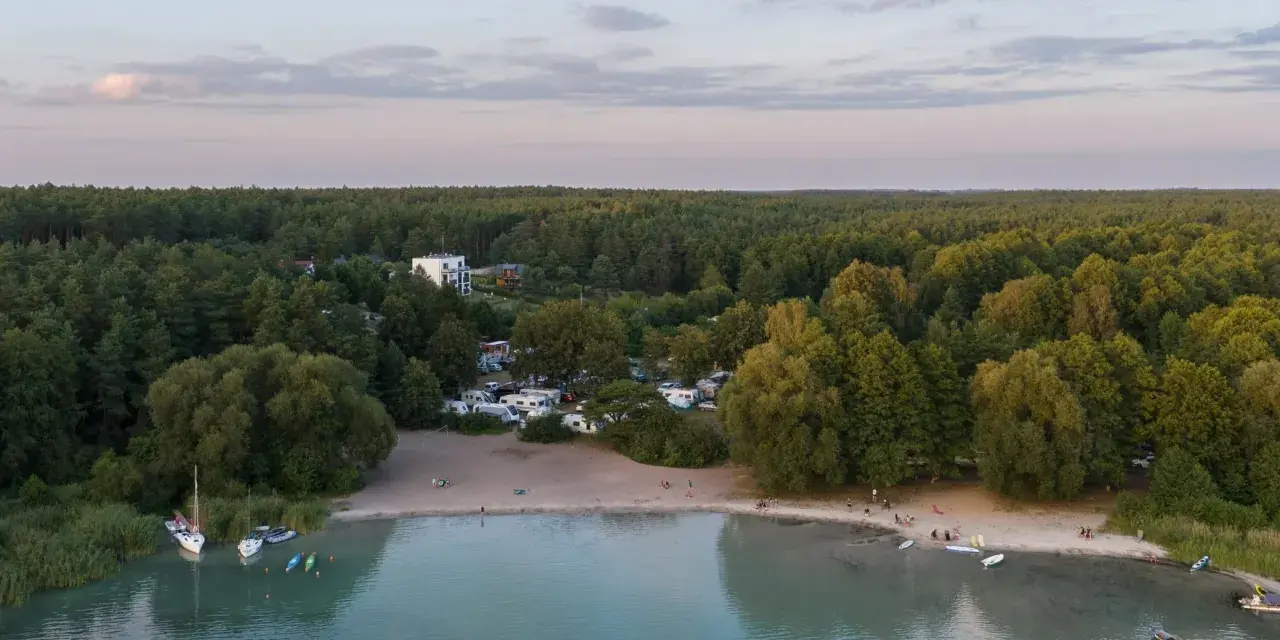 Widok na kemping nad najczystszym jeziorem w Polsce. Woda jest turkusowa, a brzeg piaszczysty. W oddali las.