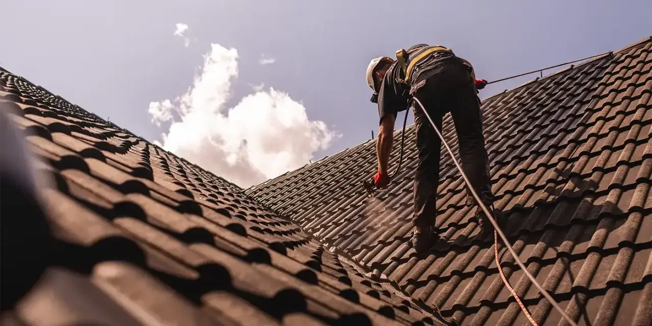 Pracownik w kasku i uprzęży zabezpieczającej wykonuje renowację dachu, natryskując dachówkę.