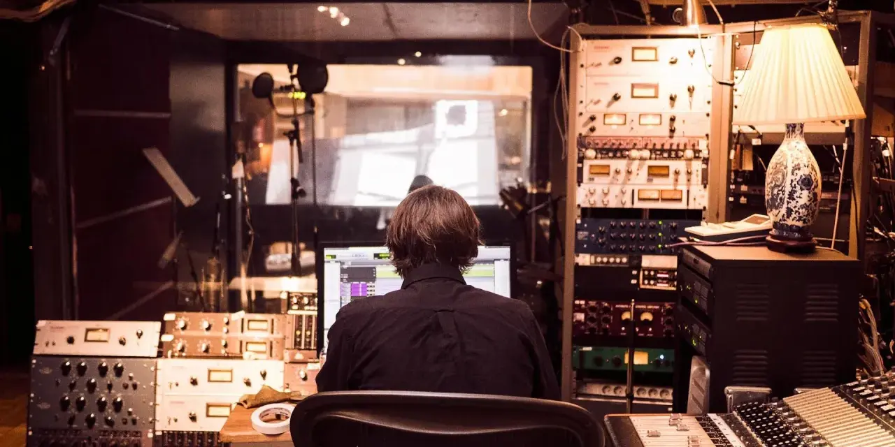 Osoba siedzi przed komputerem w profesjonalnym **recording studio**, otoczona sprzętem audio i mikserem.