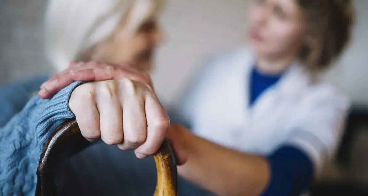 Opiekunka wspiera starszą panią, która trzyma laskę. Zastanawiasz się, ile kosztuje opiekunka z mops?