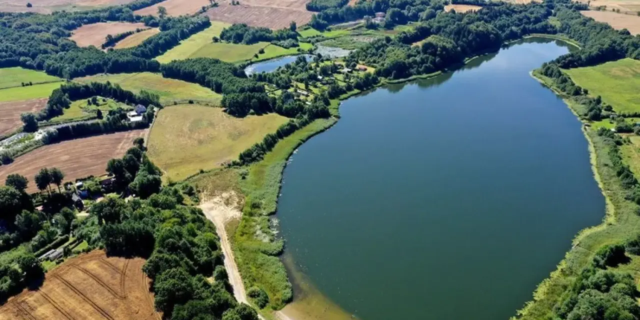 Malownicze jezioro Marszewo otoczone zielenią i polami. Widać piaszczystą plażę z pomostem i kilka samochodów.