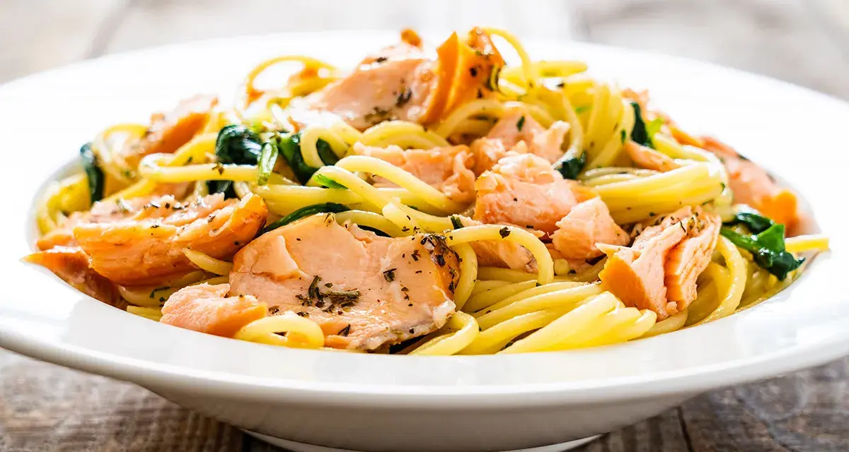 Deliciosa pasta con salmón desmenuzado y espinacas, servida en un plato blanco sobre una mesa de madera.