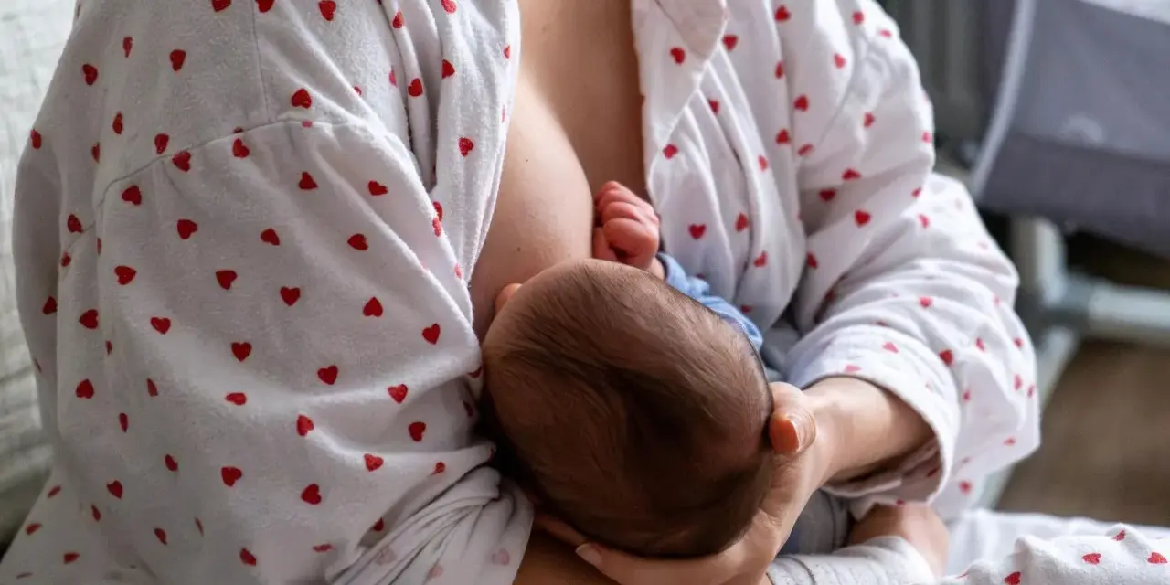 Matka karmi piersią dziecko. W tle widać łóżeczko. Zatkany kanał mleczny to częsty problem.