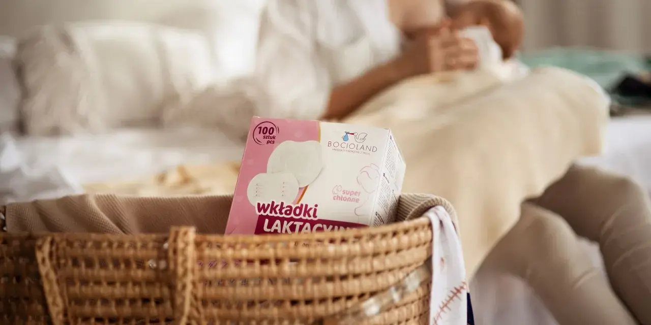 W koszyku widać opakowanie wkładek laktacyjnych Bocioland. W tle kobieta karmi piersią.