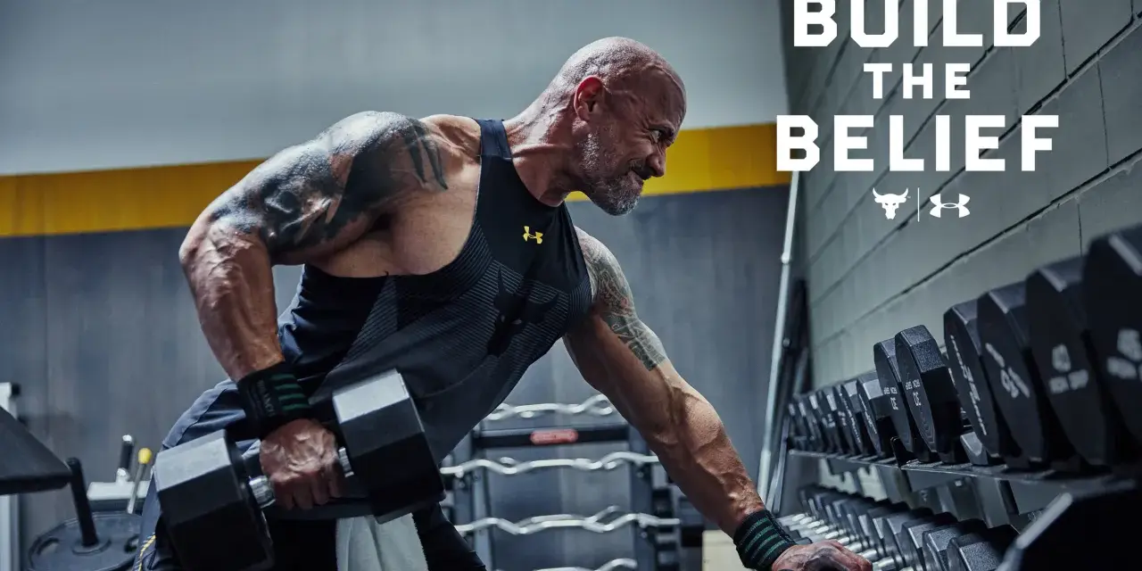 Dwayne "The Rock" Johnson w koszulce Under Armour wykonuje ćwiczenia z hantlami. W tle stojaki z ciężarami.