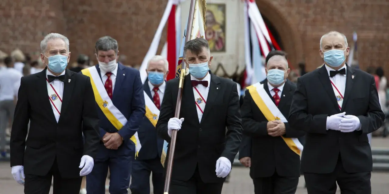 Rycerze Kolumba w garniturach i maskach maszerują z flagą.