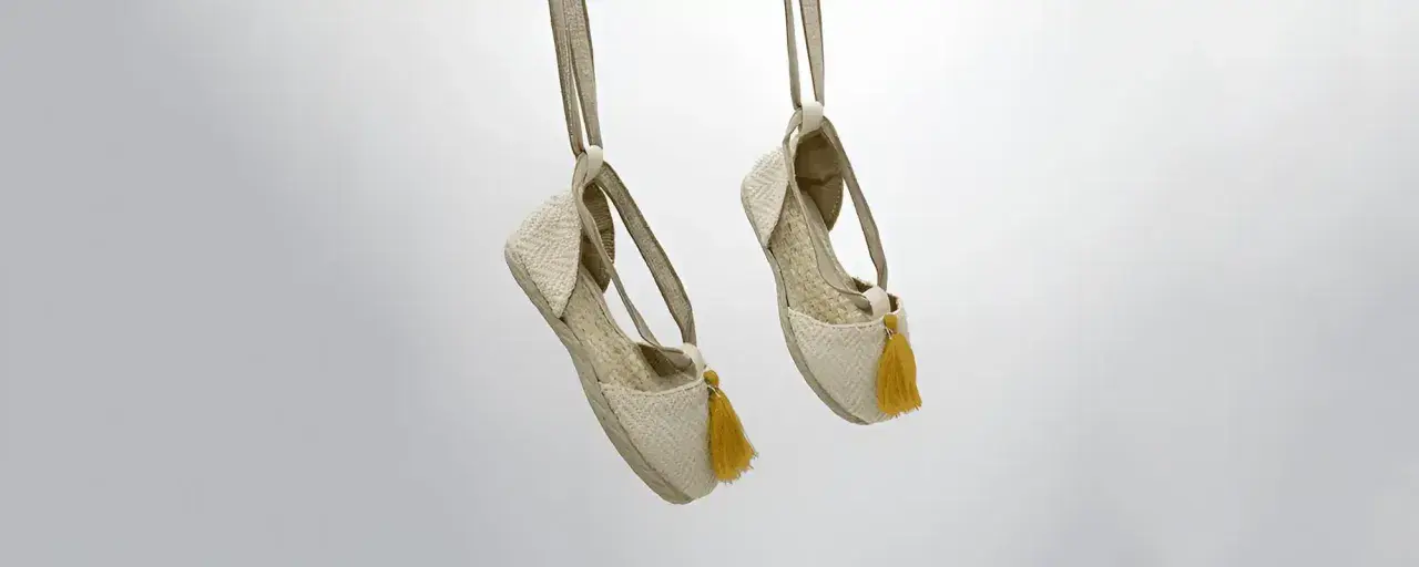 Dos alpargatas de tela beige con pompones amarillos cuelgan de tiras, mostrando su diseño veraniego.
