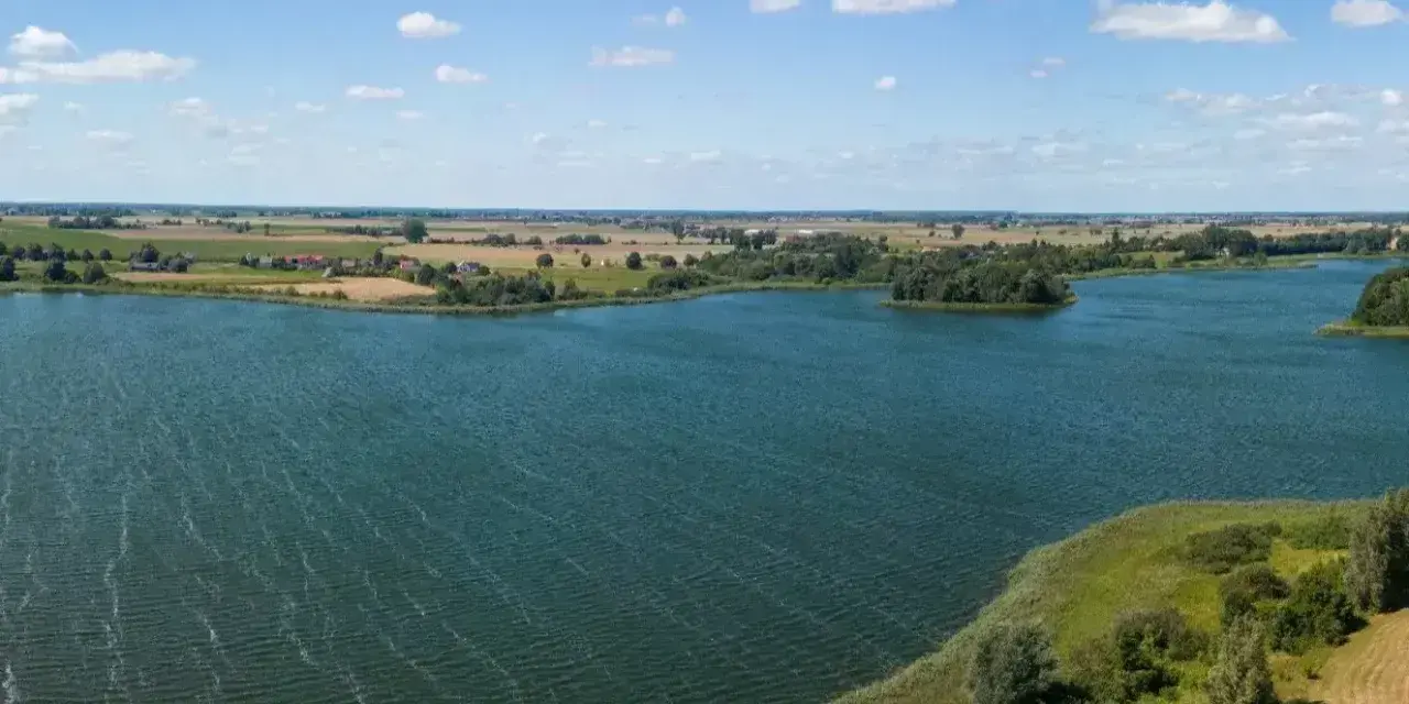 Malownicze jezioro Lednica z wyspą pośrodku, otoczone zielenią i polami. Błękitne niebo z białymi chmurami.
