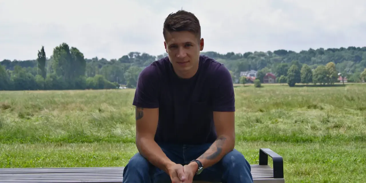 Bartłomiej Bis siedzi na ławce w parku, w tle zielone drzewa i niebo.