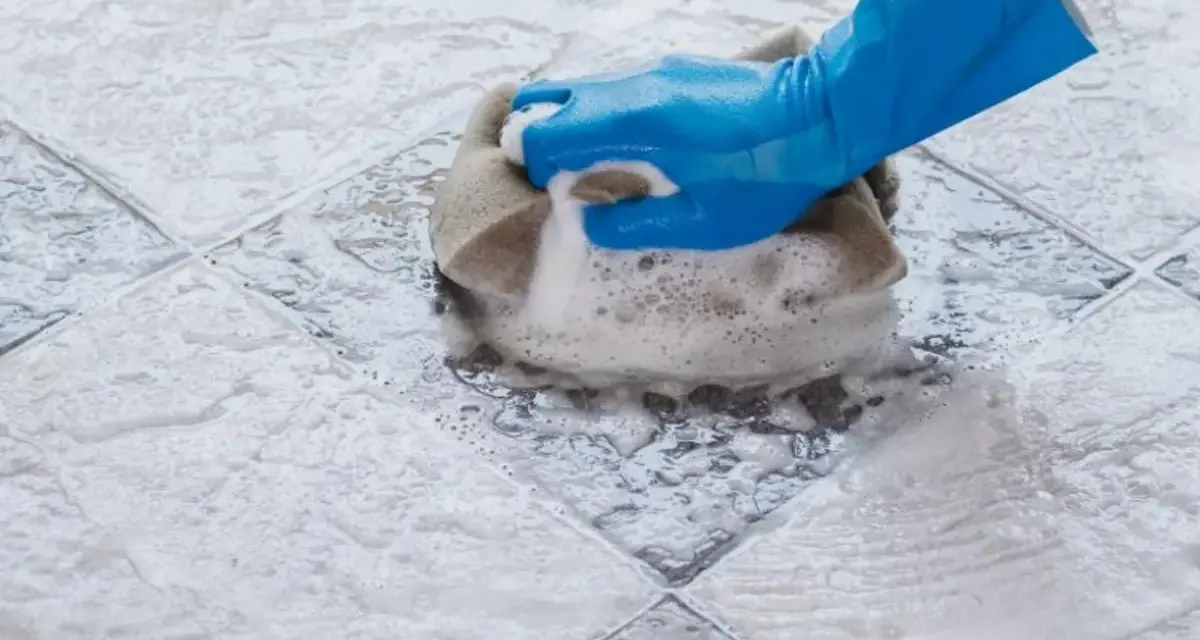 Mano con guanto blu che pulisce ceramica macchiata con spugna e schiuma.