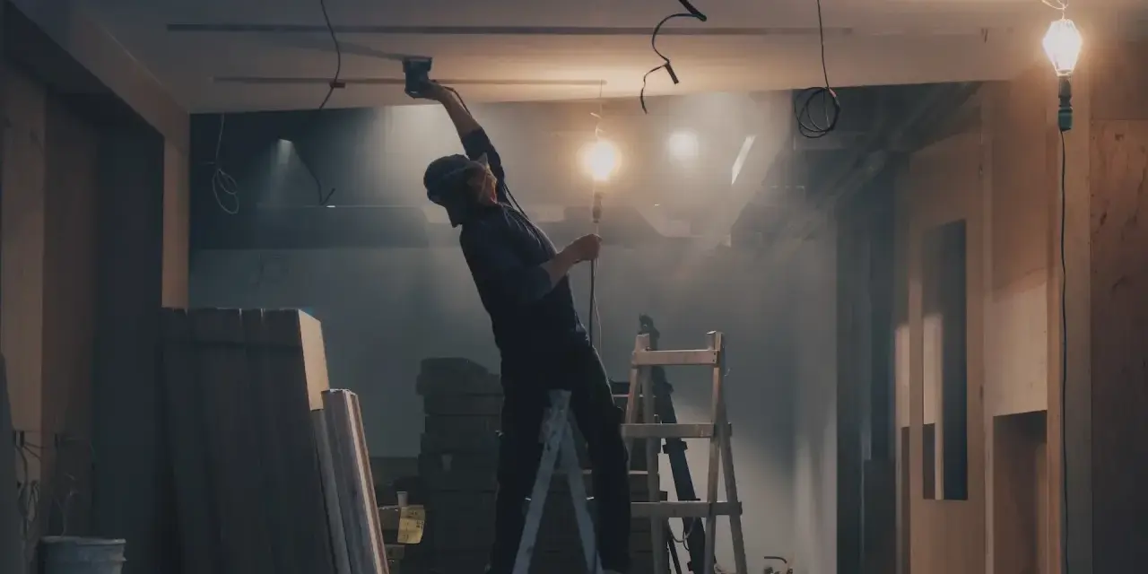 Budowlaniec na drabinie montuje sufit podwieszany, w tle widać materiały budowlane i wiszące kable.