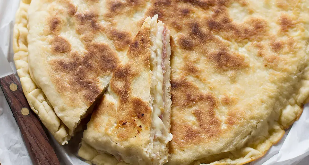 Focaccia in padella dorata e fragrante, tagliata a spicchi per mostrare il ripieno filante. Un coltello con manico in legno è appoggiato accanto.