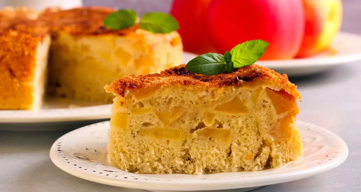 Una fetta di torta di mele vegana, soffice e ricca di mele, guarnita con menta. Sullo sfondo, una torta intera e mele rosse.