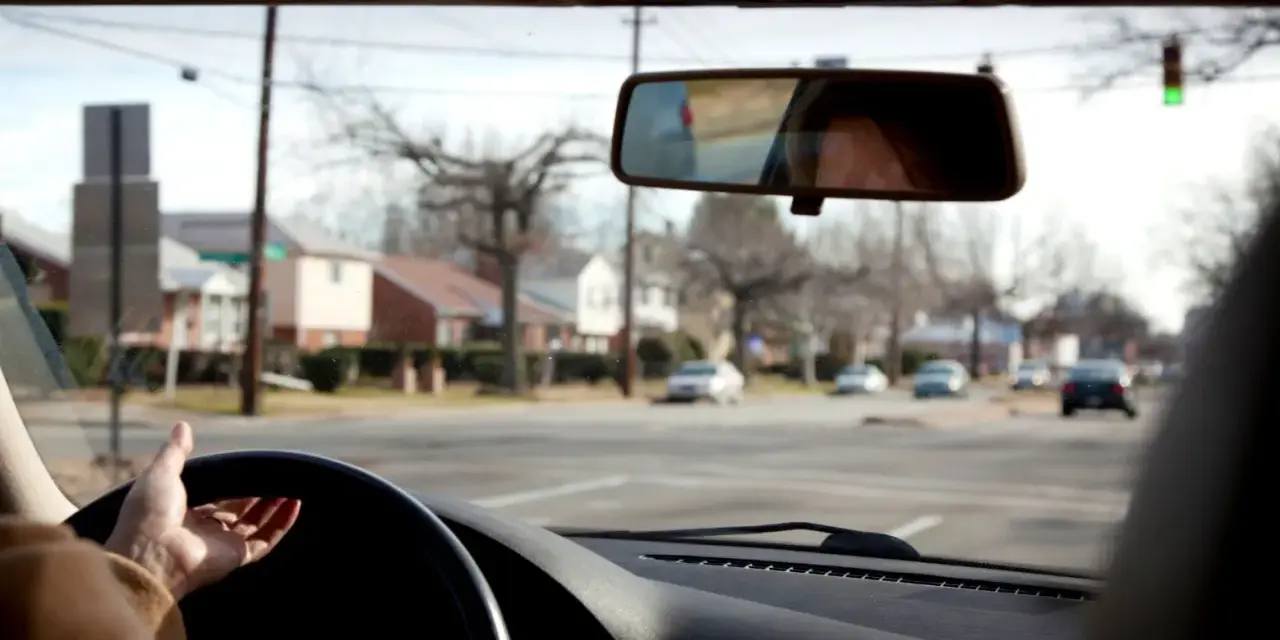 Mano sul volante, pronta per una manovra di svolta. Riflesso del volto nel retrovisore, case e alberi spogli fuori dal finestrino.
