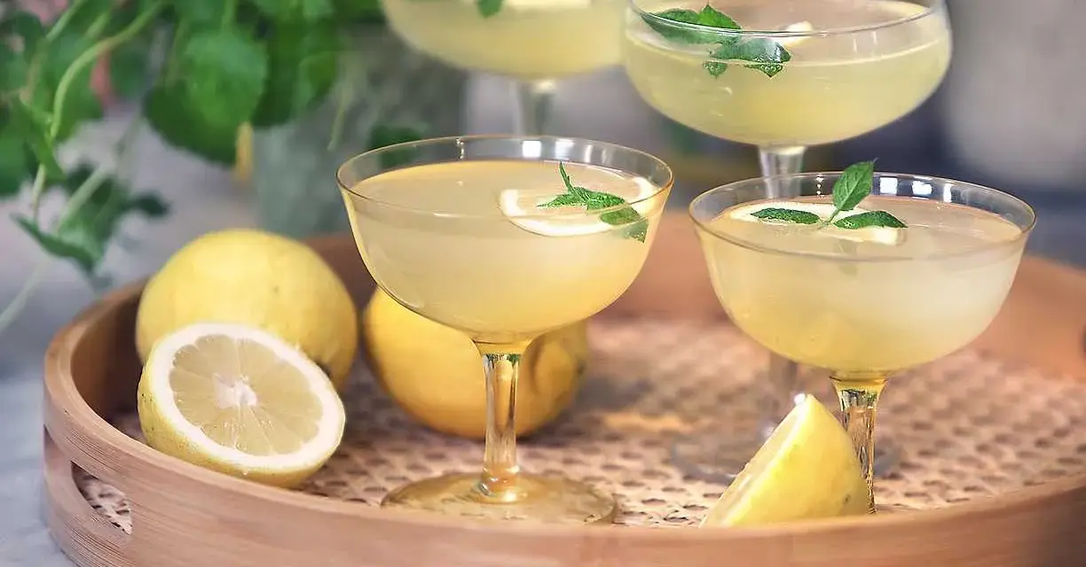 Fyra glas med en ljusgul limoncello drink, garnerade med mynta och citron. Citroner ligger på en träbricka.