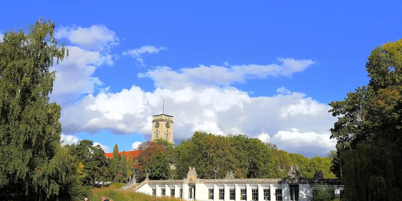 Ein sonniger Tag im **Rudolph Wilde Park**. Ein langer, weißer Pavillon spiegelt sich im Wasser, während ein Uhrenturm über Bäumen aufragt.