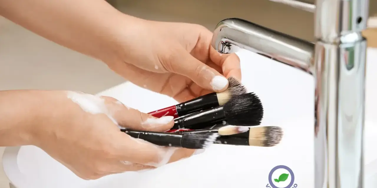 Mani che lavano pennelli da trucco insaponati sotto un rubinetto cromato. Impara come pulire i pennelli da trucco per una pelle sana.