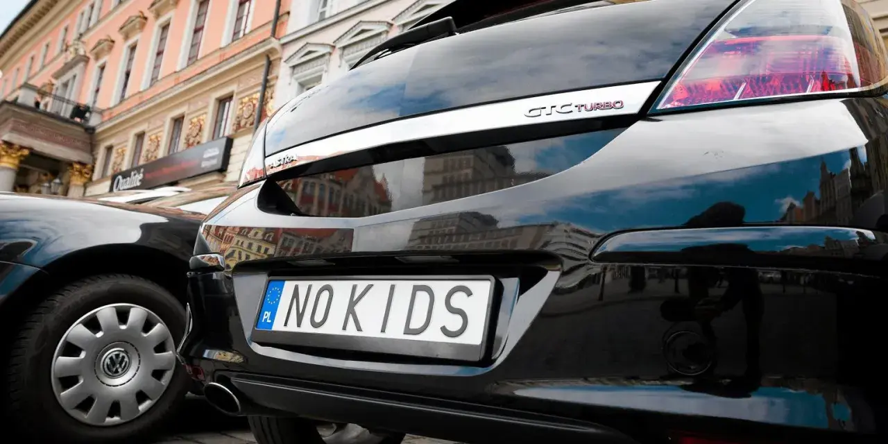 Czarne auto z indywidualnymi tablicami rejestracyjnymi "NO KIDS" na tle kamienicy.