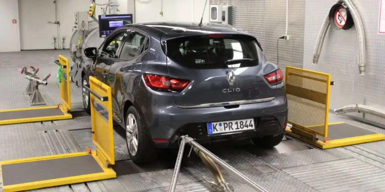 A dark grey Renault Clio is on a dynamometer in a testing facility, with exhaust pipes connected.