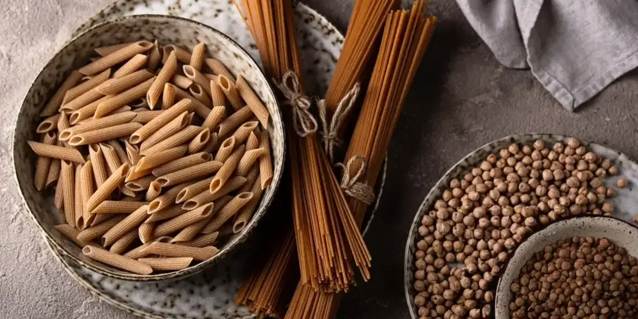 Jasny makaron penne w misce i ciemny makaron spaghetti na talerzu, obok ciecierzycy.