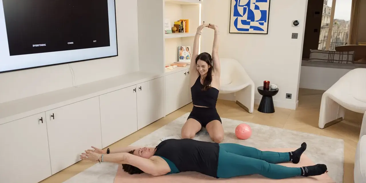 Deux femmes font du sport à la maison, une allongée sur un tapis, l'autre assise. Idéal pour un sport canicule sans sortir.