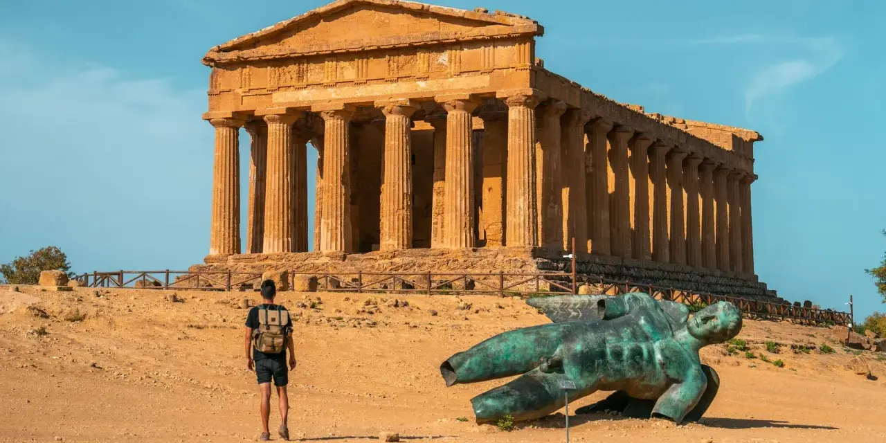 A lone traveler explores the Valley of the Temples in Sicily, standing before an ancient Greek temple and a fallen bronze statue.