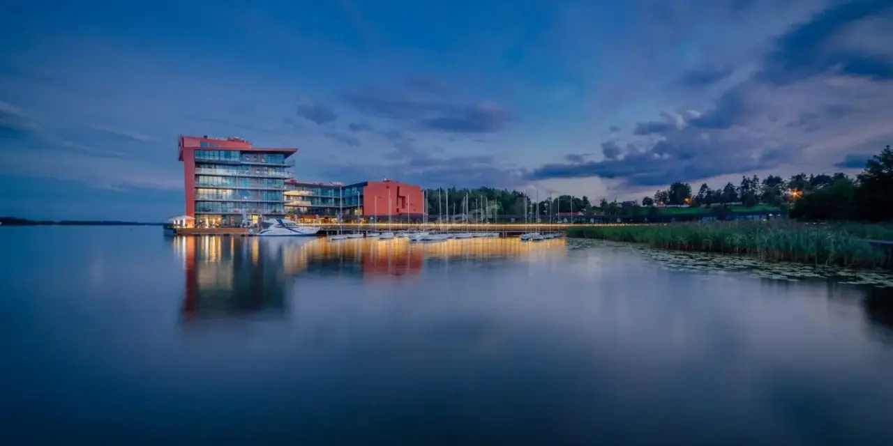 Nowoczesny hotel na ptasiej wyspie, otoczony spokojną wodą i zielenią, z łodziami przy pomoście.