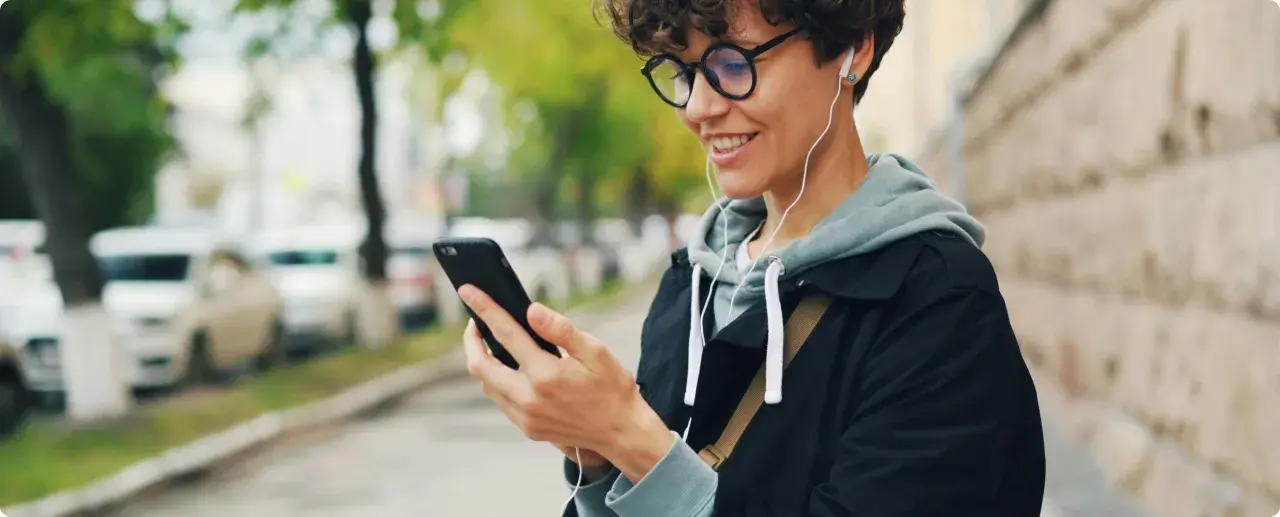 Uśmiechnięta osoba w słuchawkach patrzy na telefon. Czy wiesz, co to jest impedancja w słuchawkach i jak wpływa na dźwięk?