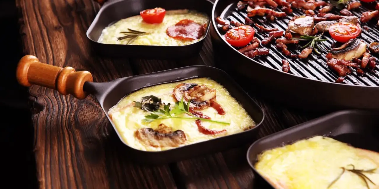 Raclette co to? Roztopiony ser z dodatkami w małych patelenkach i grillowane mięso z pomidorkami na drewnianym stole.