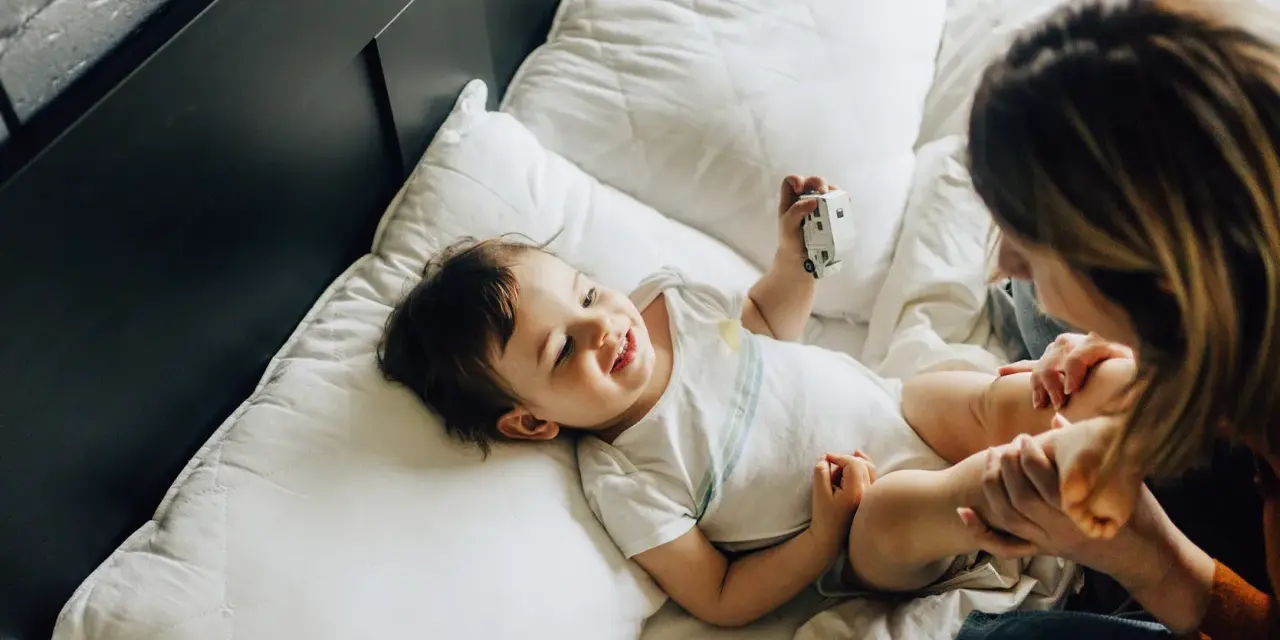 Uśmiechnięte dziecko bawi się zabawką na łóżku. Mama patrzy na malucha. Co to za mastka?