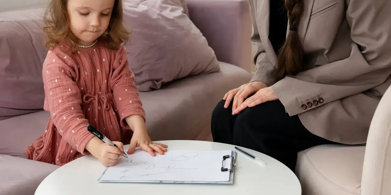 Mała dziewczynka w różowej sukience rysuje na kartce, podczas gdy dorosła osoba w beżowej marynarce obserwuje. Sesja ados 2.