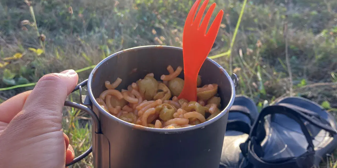 Un repas simple pour savoir que manger en randonnée : des pâtes avec des olives dans une tasse de camping, avec une fourchette orange.