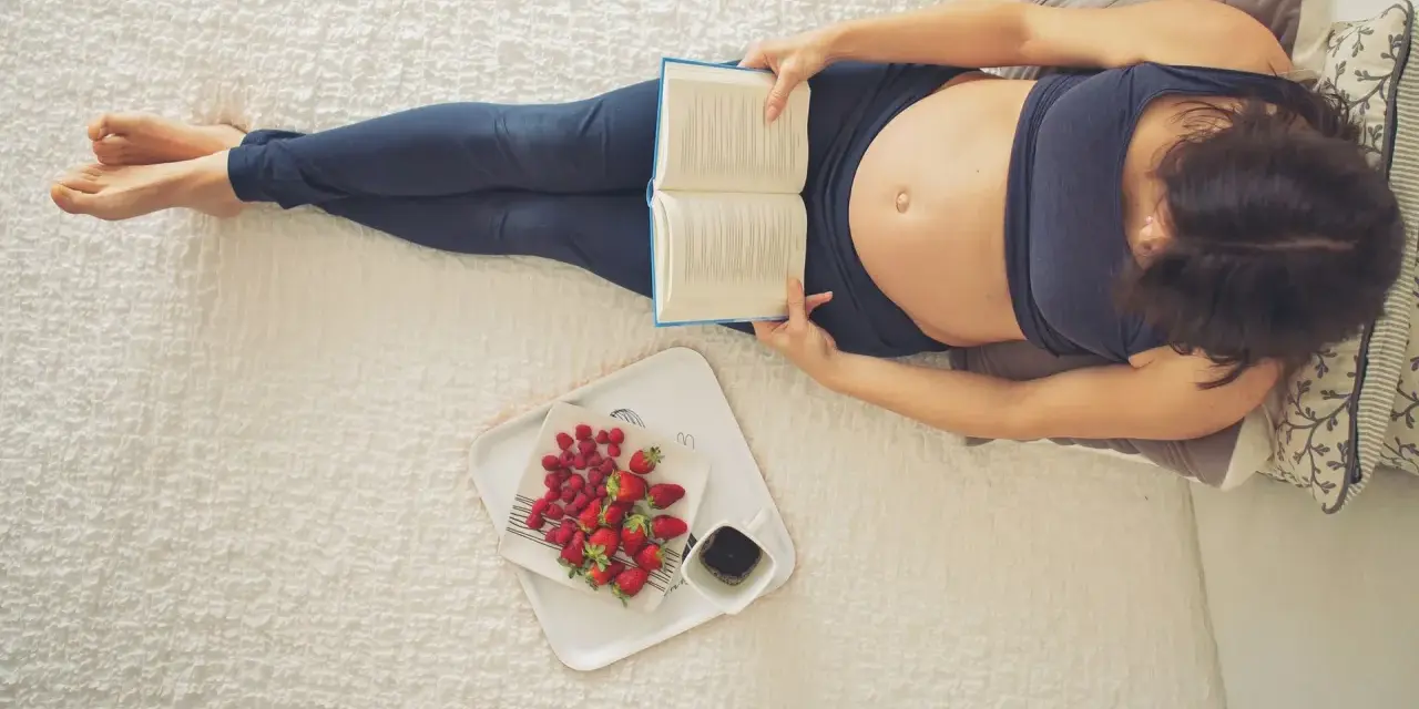 Kobieta w 22 tygodniu ciąży, czyli w 5. miesiącu, relaksuje się z książką i owocami.