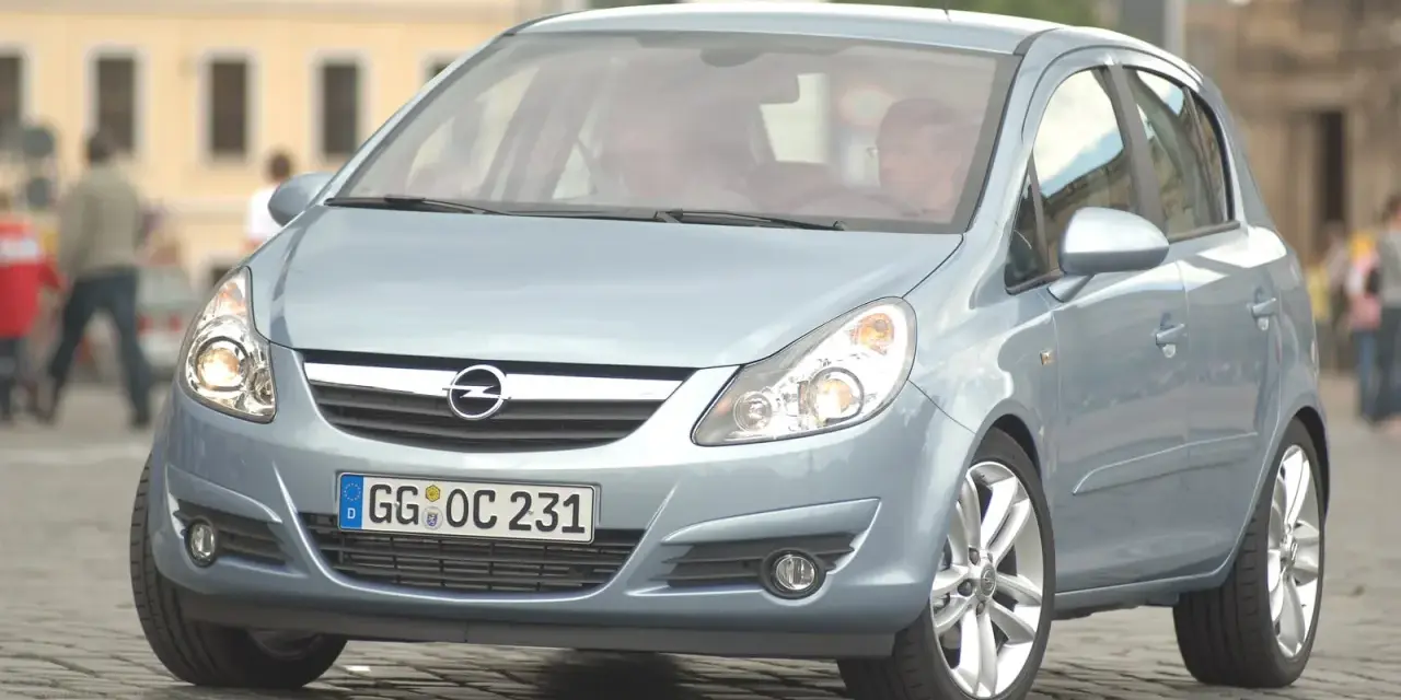 Jasnoniebieski Opel Corsa D z włączonymi żarówkami z przodu, zaparkowany na bruku.