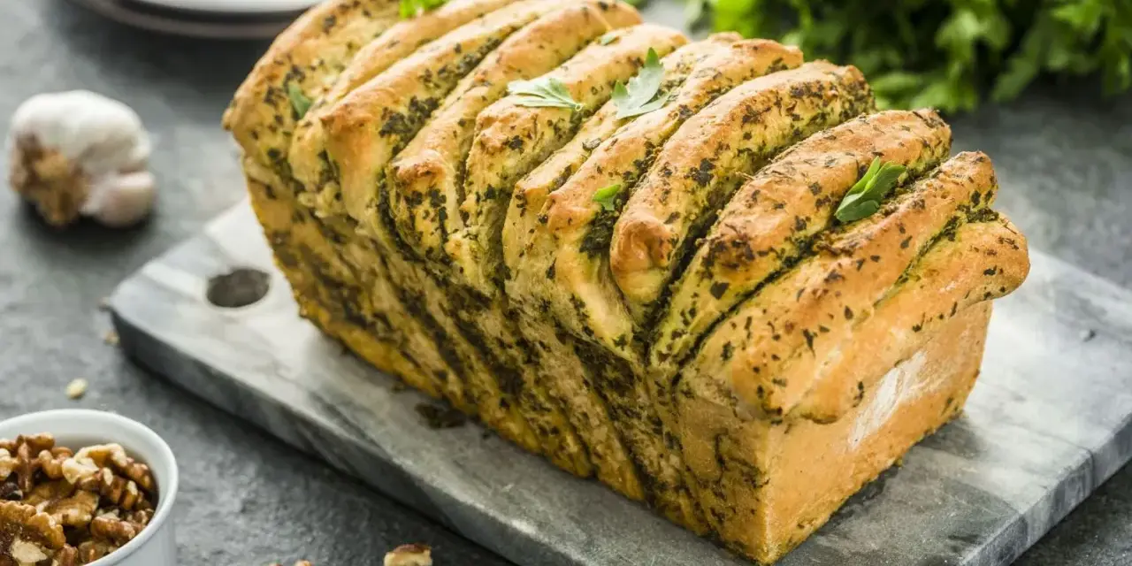 Pyszny chlebek rwany z pesto, udekorowany listkami bazylii, prezentuje się apetycznie na kamiennej desce. Obok czosnek i orzechy włoskie.