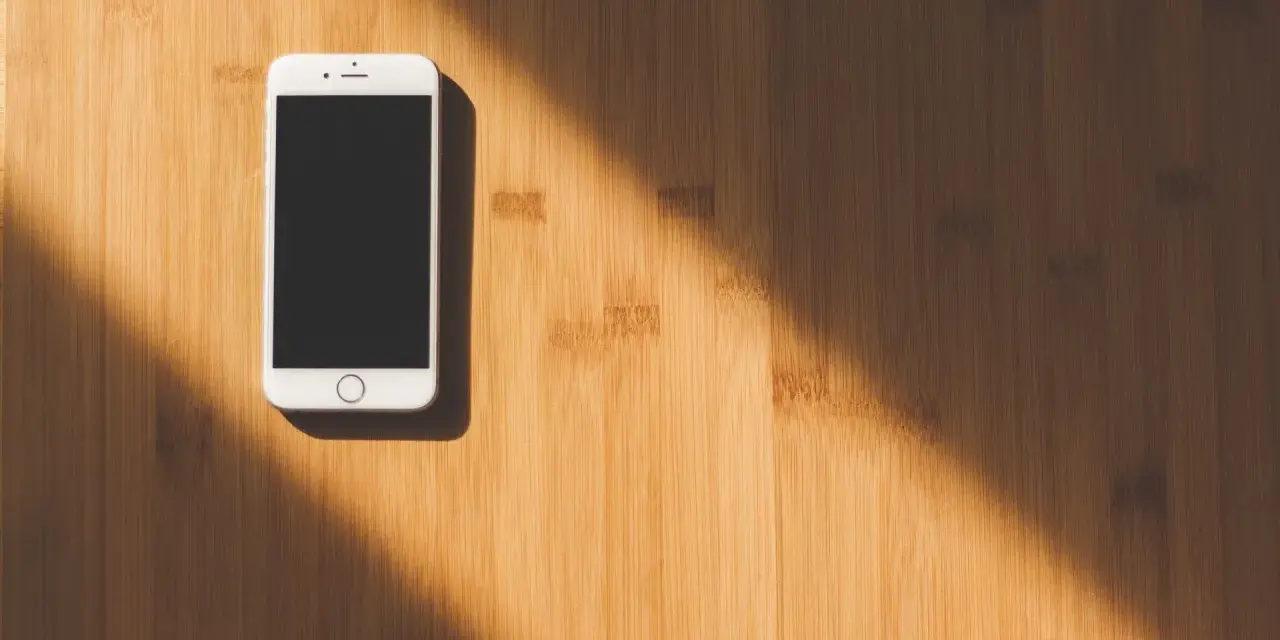 Biały smartfon leży na drewnianym blacie. Słońce rzuca cień. Jak namierzyć telefon, gdy zaginie?