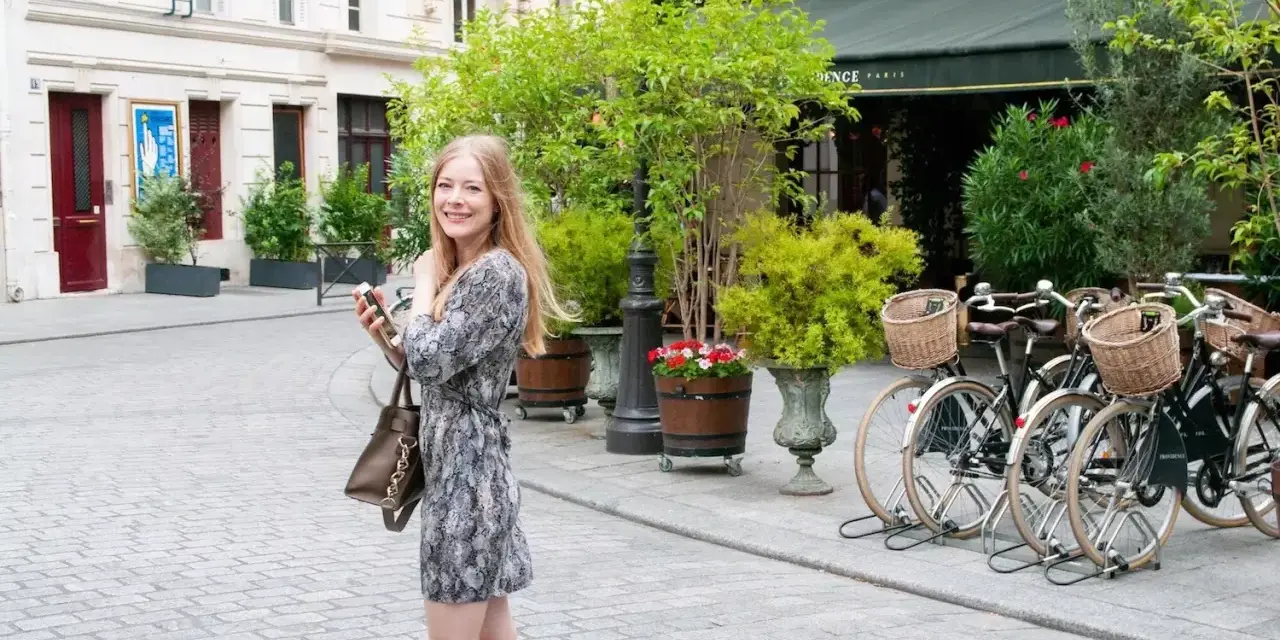 Uśmiechnięta kobieta z telefonem w ręku spaceruje po Paryżu. W tle widać rowery i urokliwe kamienice, jakby z piosenki o Paryżu.