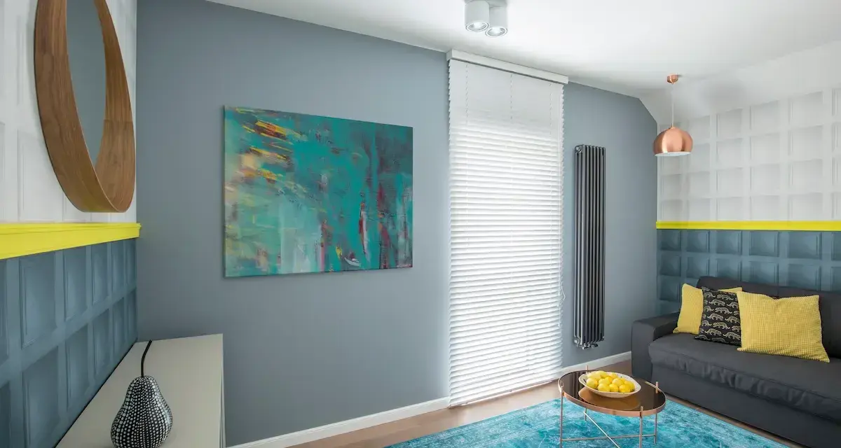 Sala de estar con sofá gris, alfombra turquesa y arte abstracto en tonos fríos. Un espejo redondo de madera y detalles en amarillo complementan el espacio.