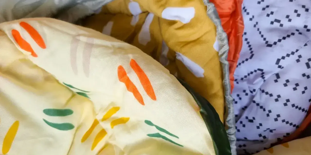Bunte Muster auf einem Schlafsack aus Baumwolle oder Polyester. Ein gelber Stoff mit orangen und grünen Streifen, daneben ein orangefarbener und ein weißer Stoff mit schwarzen Punkten.