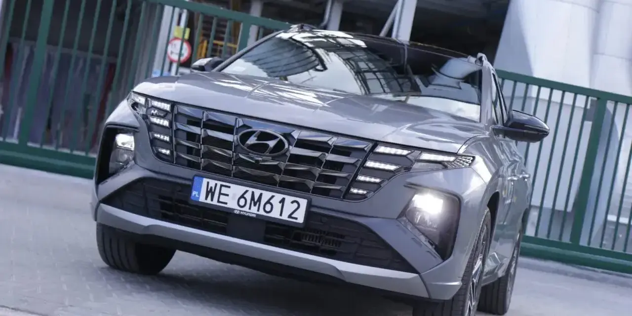 Szary Hyundai Tucson hybryda z charakterystycznym grillem i światłami LED, zaparkowany przed zielonym ogrodzeniem.