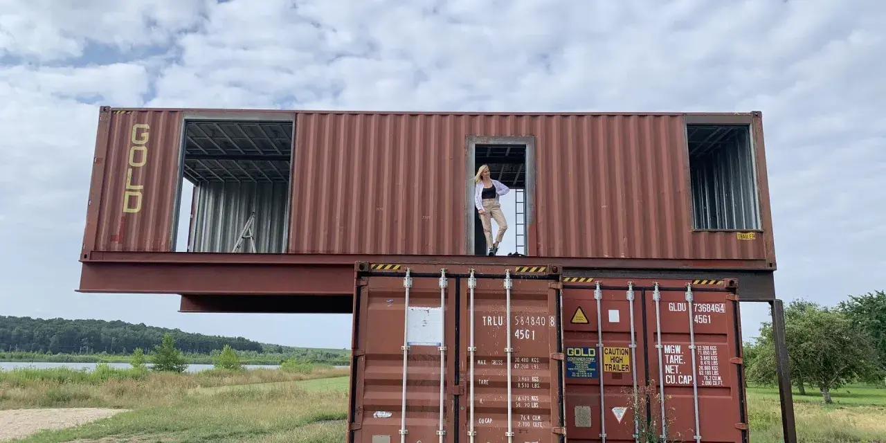 Kobieta stoi w drzwiach domu z kontenera, który jest częścią dwupoziomowej konstrukcji.