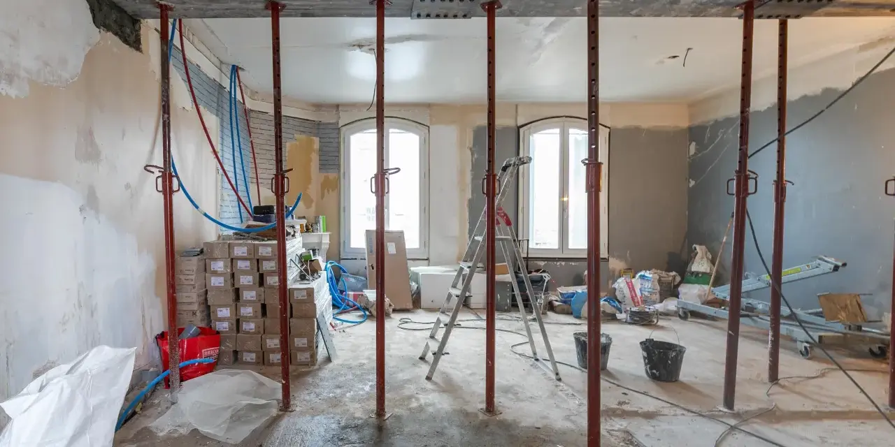 Chantier d'une maison phenix 1970 : poutres métalliques, étais, tuyaux et matériaux de construction dans une pièce en rénovation.