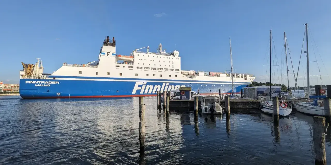 Duży prom Finnlines "Finntrader Malmö" cumuje w porcie, gotowy do rejsu. W tle widać żaglówki. Prom do Finlandii czeka na pasażerów.