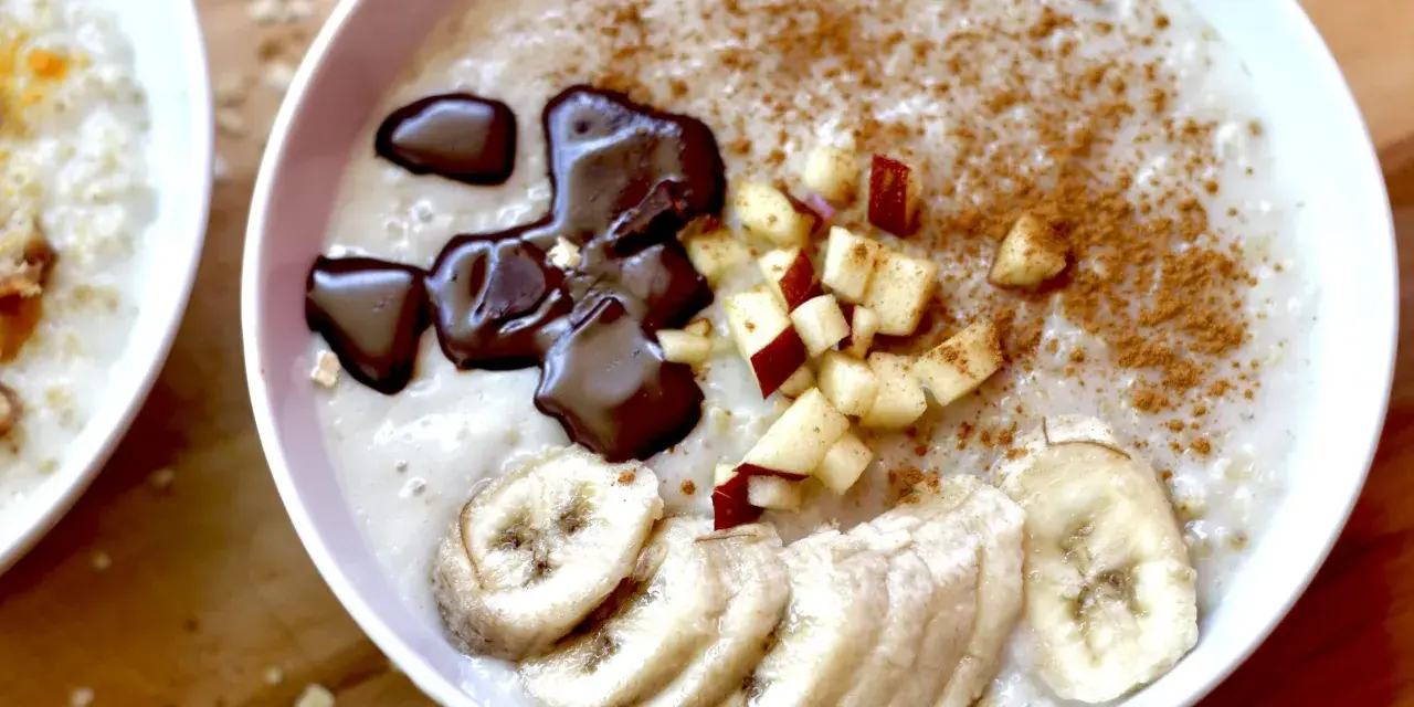 Un tazón de avena cremosa, perfecto para aprender como hacer porridge. Decorado con rodajas de plátano, trocitos de manzana, canela y un toque de chocolate derretido.
